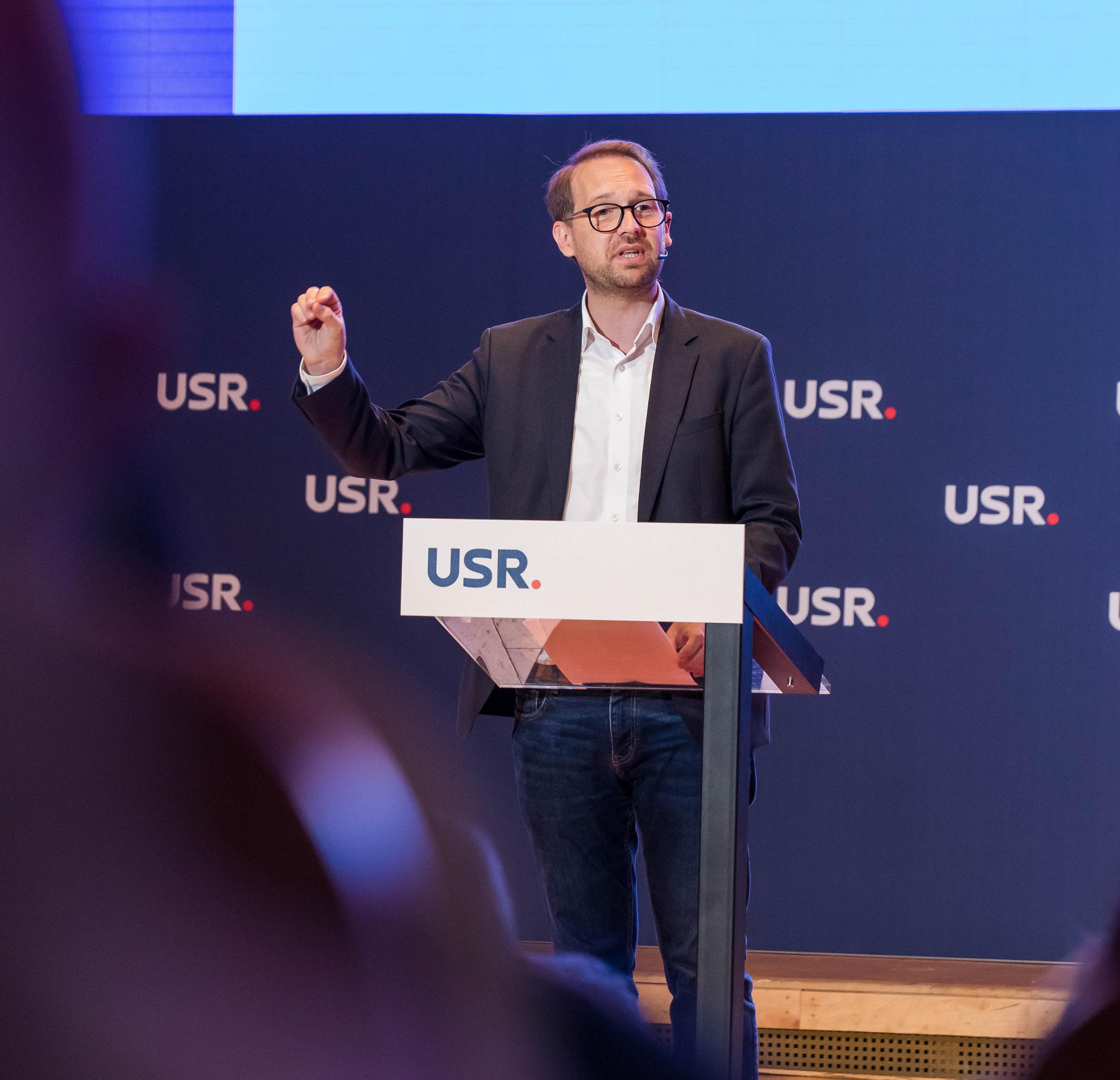Dominic Fritz vorbind la un pupitru cu sigla USR, cu o mână ridicată, în fața unui panou cu sigle USR