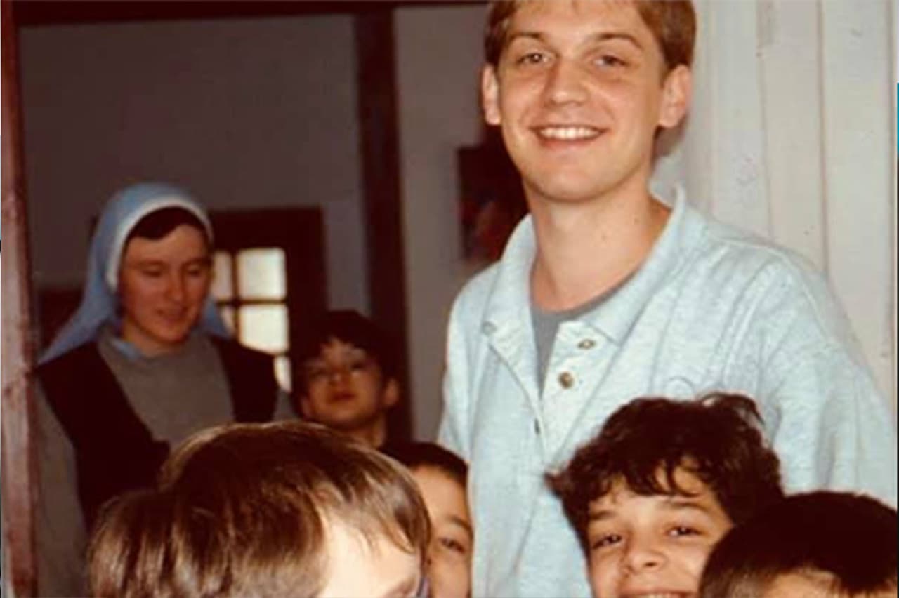 Dominic Fritz as a young volunteer, smiling among the children of a home in Timișoara, with a nun standing in the doorway behind him
