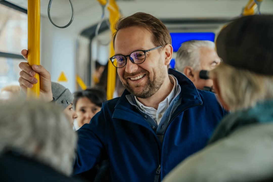 Dominic Fritz holding on to a handrail on a Timișoara public transport vehicle, talking to the passengers around him